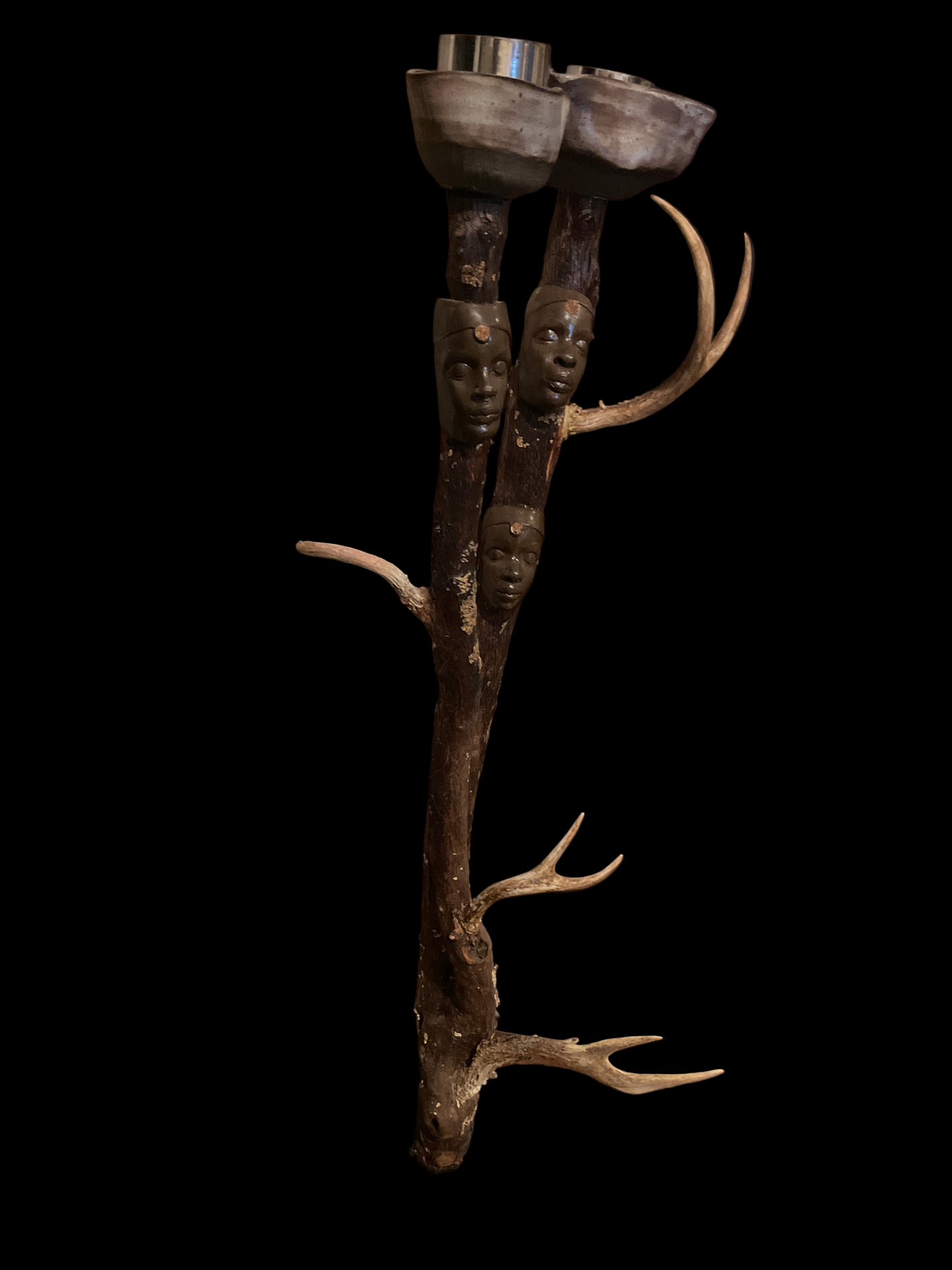 This ancestral candle holder honors the sacred power of our ancestors woven through nature. Two wheel-thrown vessels cradle the light of remembrance, while the faces of African queens emerge from the limbs of the huckleberry tree. its spirit is carried through their expression. Adorned with deer antlers, symbols of intuition, protection, and noble strength, the sculpture becomes an altar of lineage. It invites reflection , and reverence for the ancient wisdom of our ancestors.