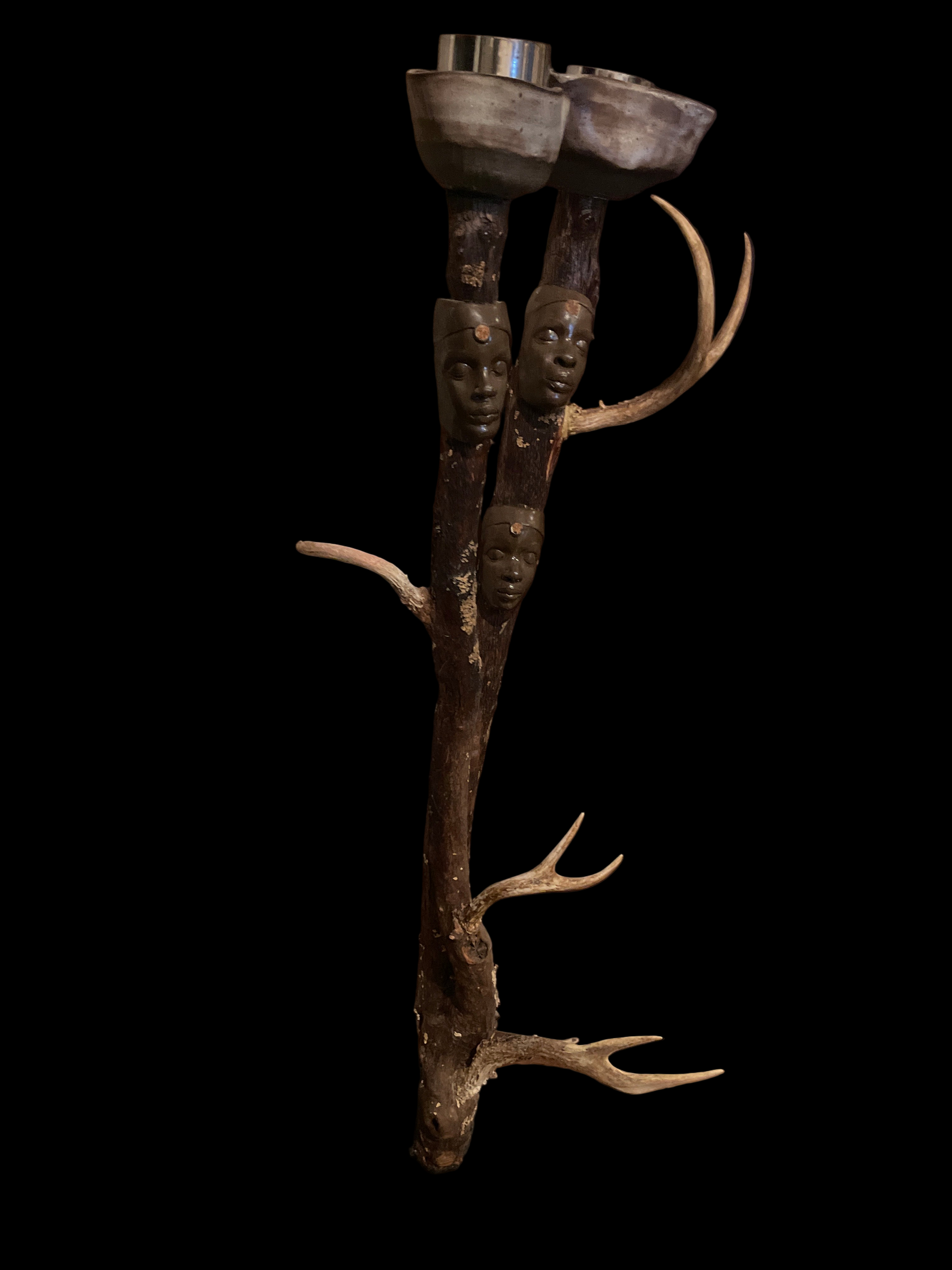 This ancestral candle holder honors the sacred power of our ancestors woven through nature. Two wheel-thrown vessels cradle the light of remembrance, while the faces of African queens emerge from the limbs of the huckleberry tree. its spirit  is carried through their expression. Adorned with deer antlers, symbols of intuition, protection, and noble strength, the sculpture becomes an altar of lineage. It invites reflection , and reverence for the ancient wisdom of our ancestors. 
